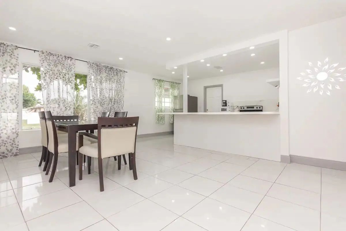 Bright open dining room and kitchen with white tile floors, dark table and chairs, and large windows.