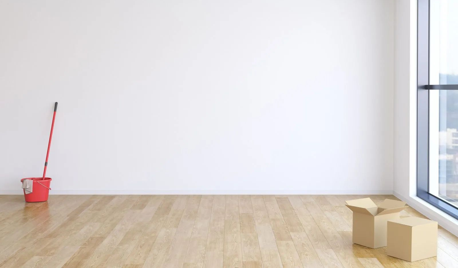 Empty white room with wood floor, a red mop and bucket on the left, and moving boxes by a window on the right
