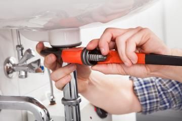 A person uses a red pipe wrench to tighten a metal drain pipe under a white sink.