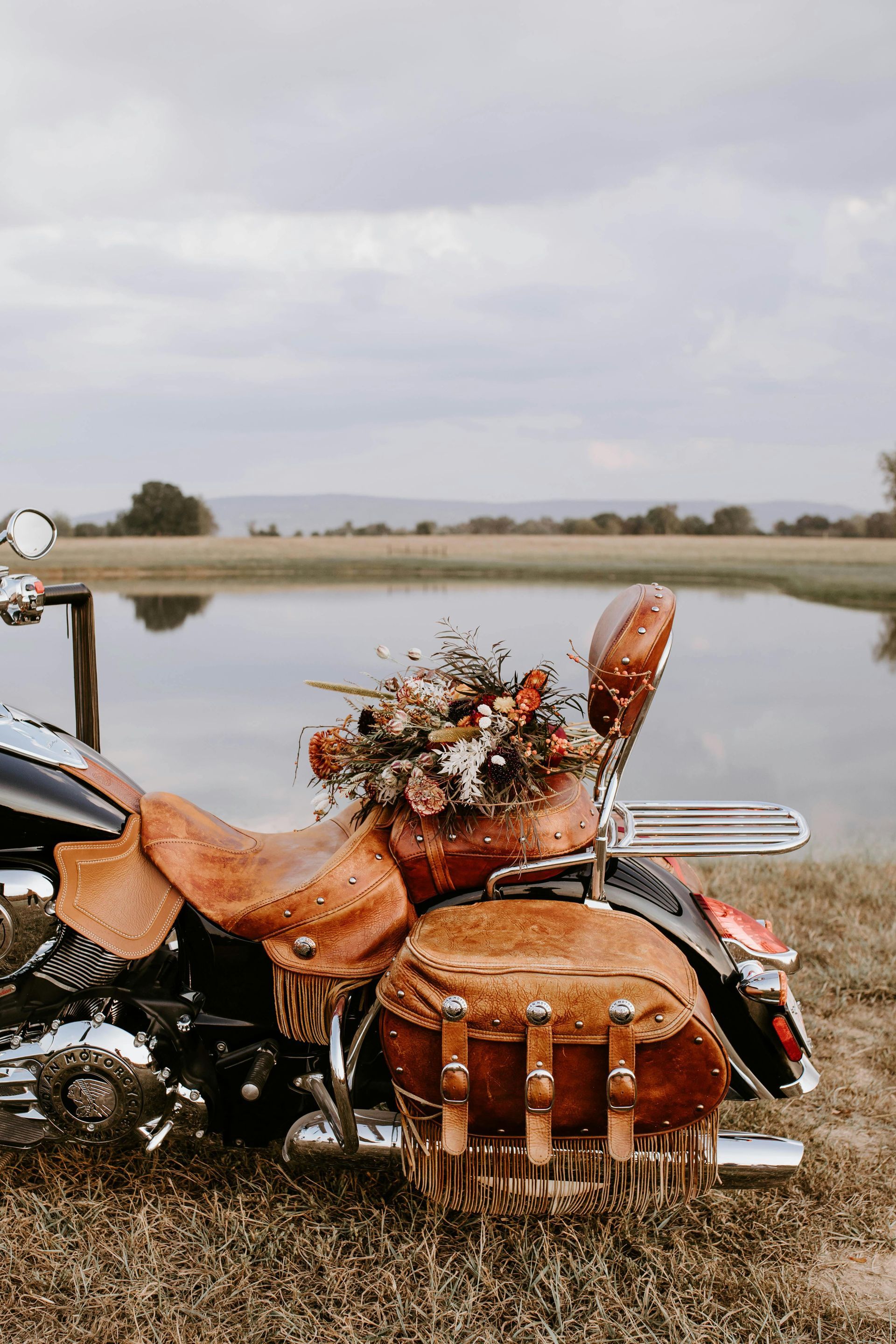 A motorcycle is parked in the grass next to a body of water with a fall bouquet on the back seat.