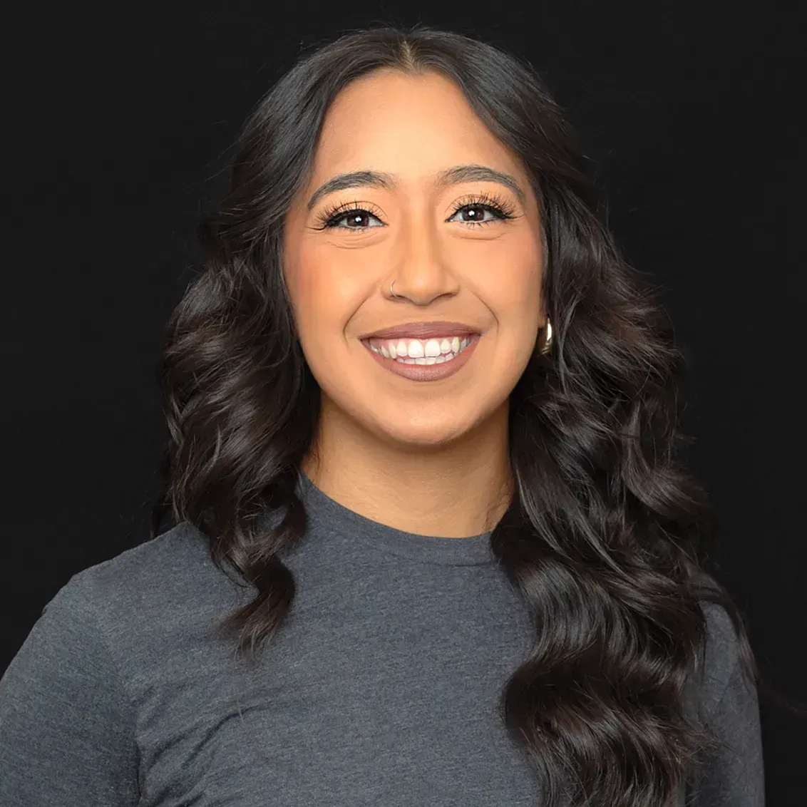 Smiling woman with long, dark wavy hair, wearing a gray shirt, and gold earrings, against a black background.