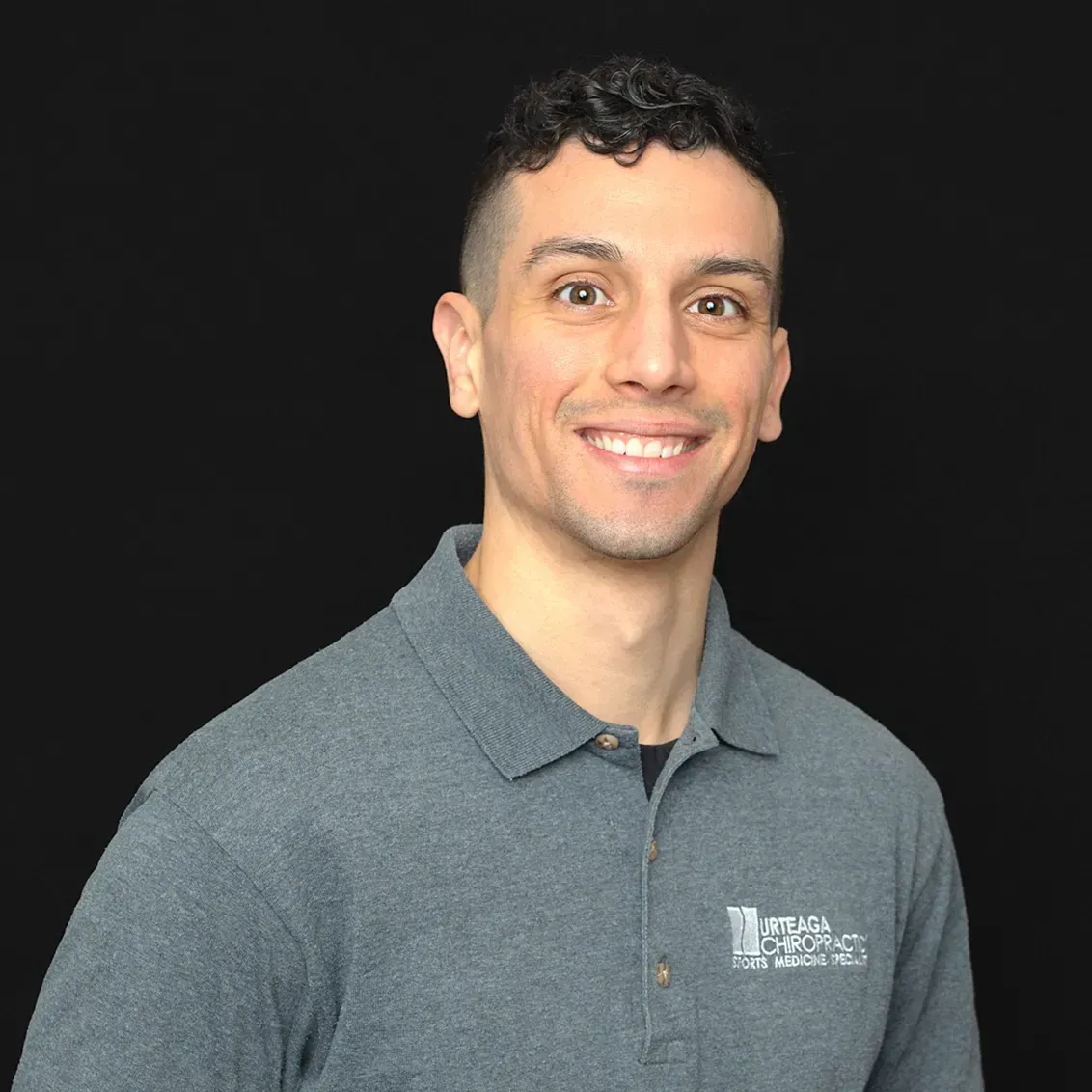 Man smiling, wearing a gray polo shirt, dark hair, against a black backdrop.