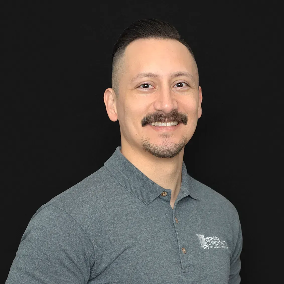 Man with a mustache, wearing a gray polo shirt, smiles against a black background.