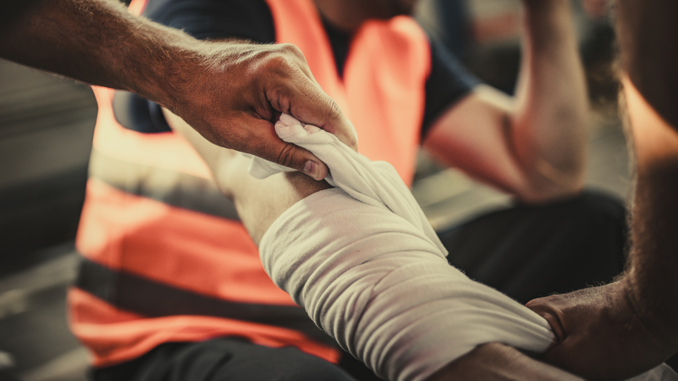 Person wrapping bandage around another's arm, outdoors. The person helping wears an orange safety vest.