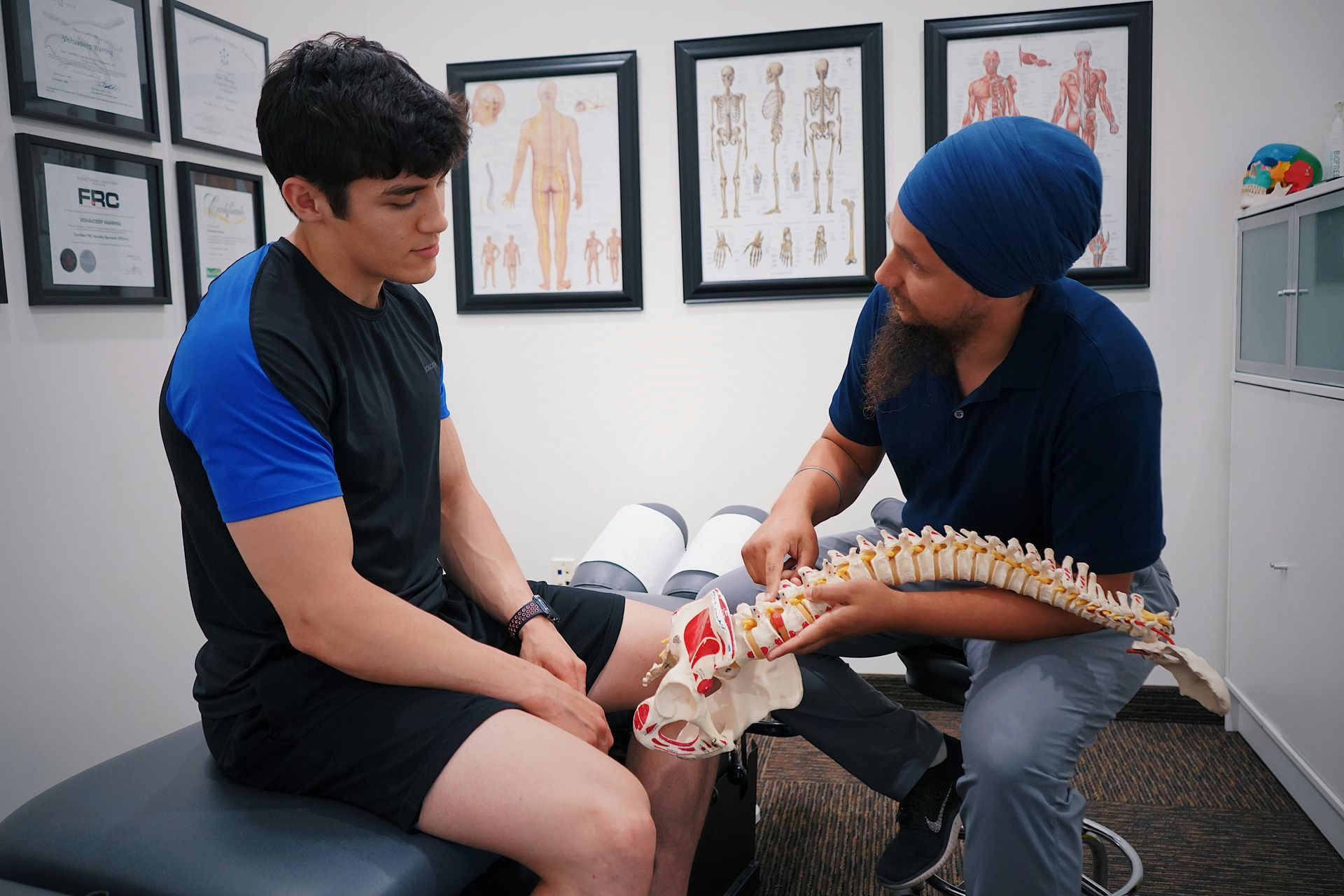 Chiropractor examining a seated patient’s lower back with a spine model in a clinic room