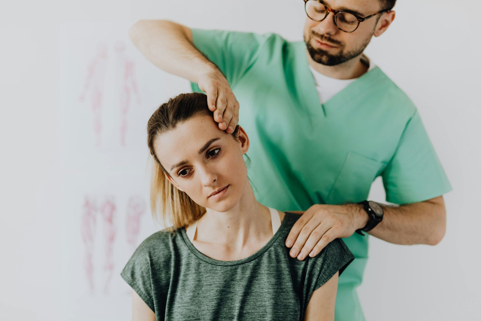 Chiropractor adjusting woman's neck, green scrubs, white wall backdrop.