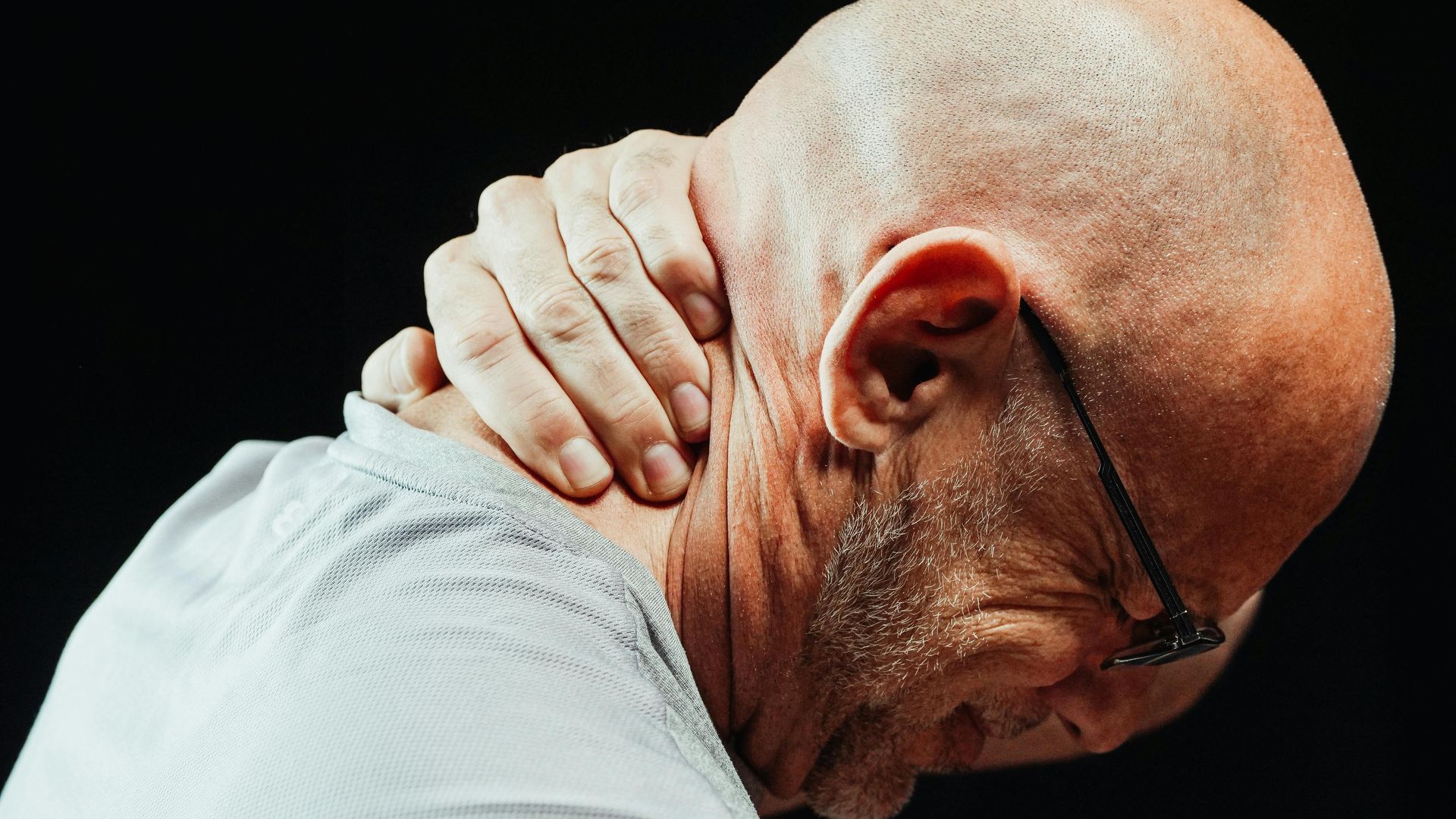 A person with a shaved head in a light shirt holding the back of their neck with an expression of physical pain.