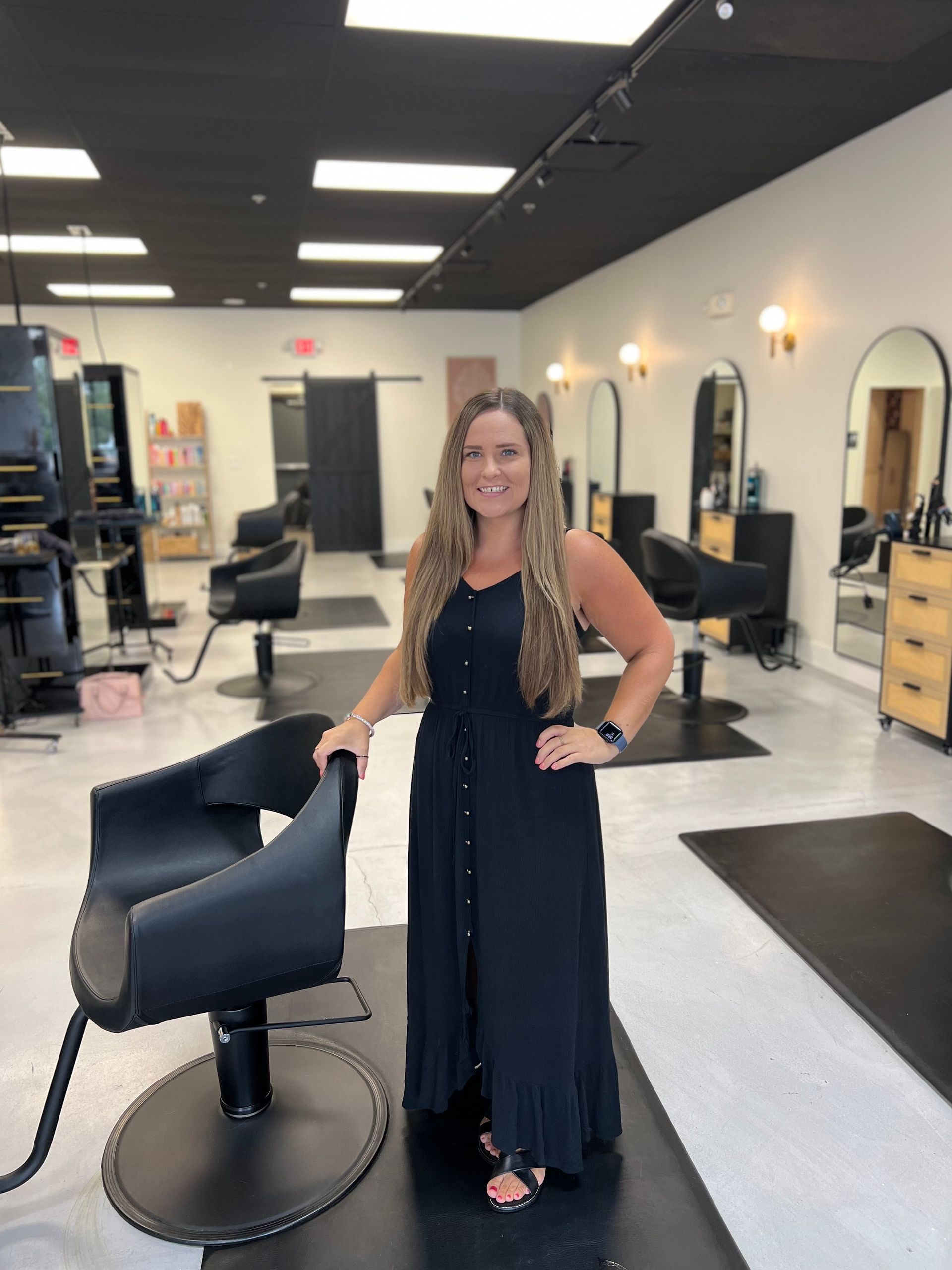 A woman in a black dress is standing next to a chair in a salon