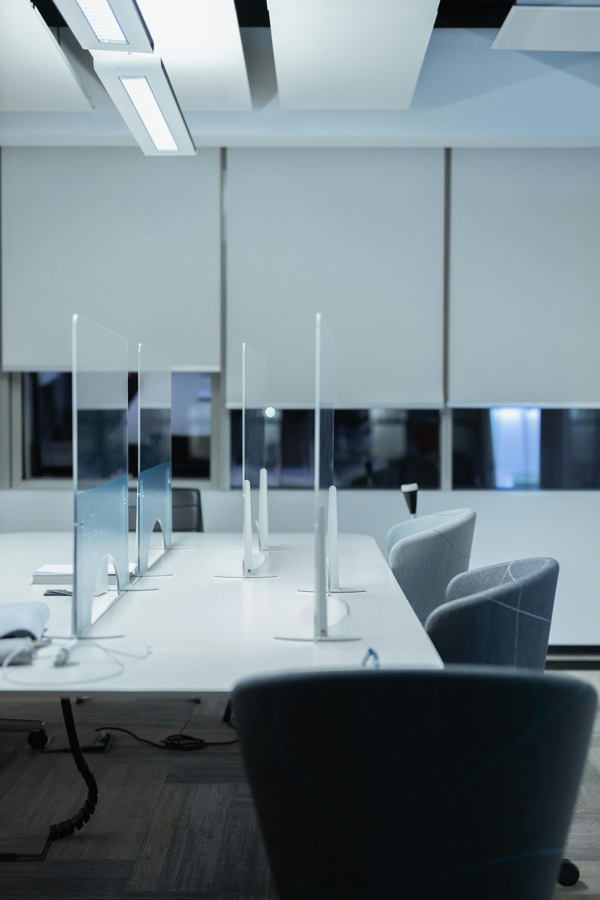 Office desks with dividers, monitors, and chairs. White surfaces, neutral tones, and window light.