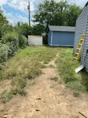 A dirt path leads through a grassy backyard toward a blue storage shed and a smaller, white wooden structure.