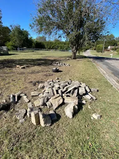 A pile of gray stones and construction debris sits on a grassy lawn near a roadside under a large tree.