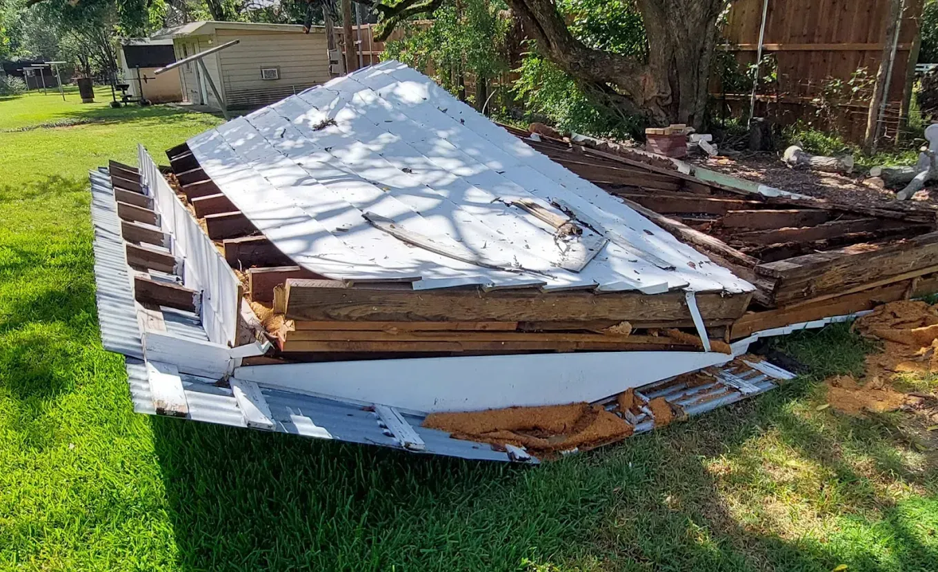 A collapsed roof section with exposed wooden joists and white panels lying on a grassy lawn.