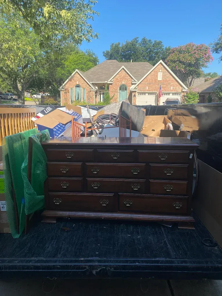 A wooden dresser with drawers sits in the back of a truck, with a brick house visible in the background.