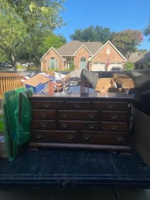 A dark wood dresser sits in the bed of a pickup truck, parked in front of a residential brick house on a sunny day.