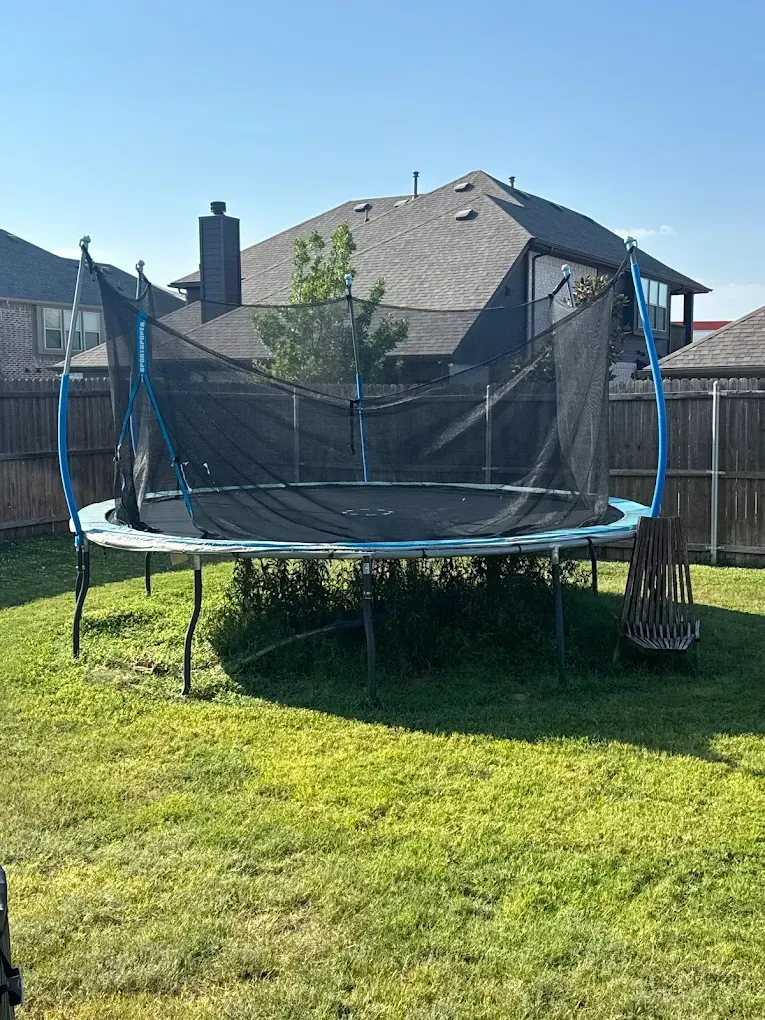 A large, round backyard trampoline with a black safety net enclosure, situated on a grassy lawn near a wooden fence.