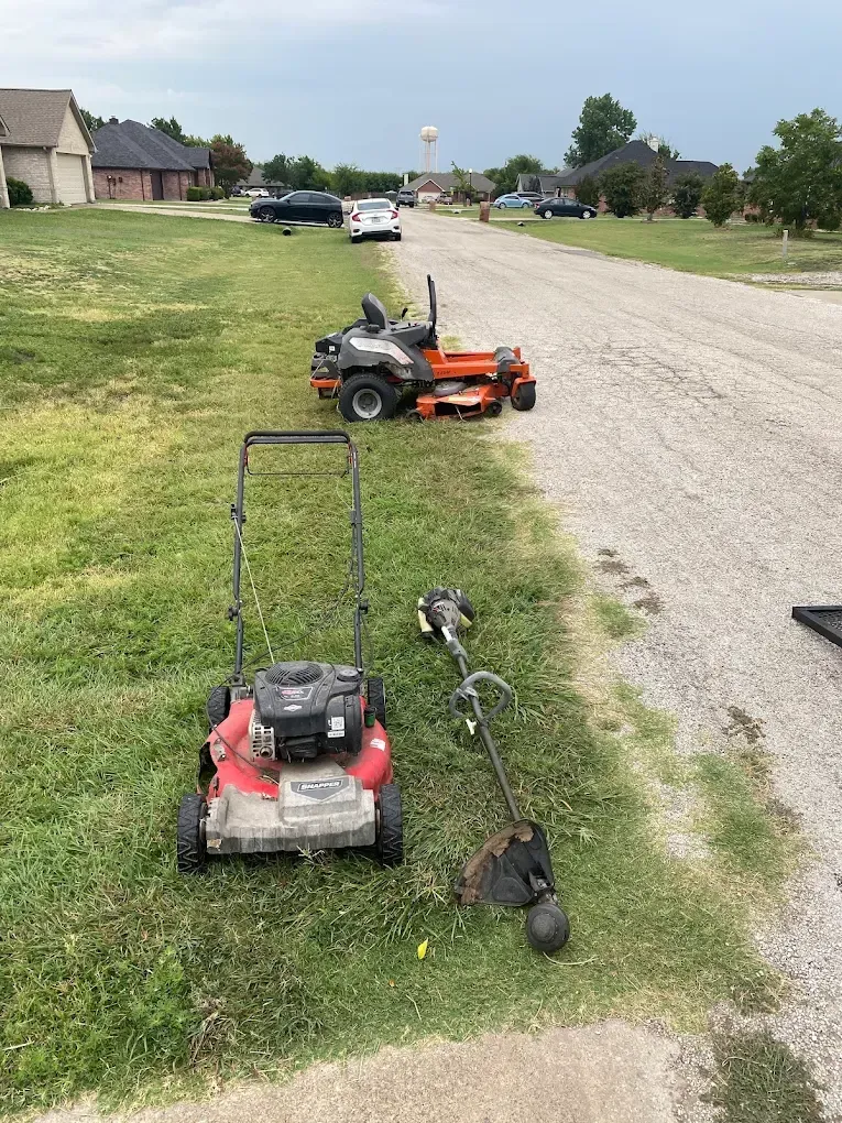 A red lawnmower, a string trimmer, and an orange zero-turn mower are lined up on the grass beside a gravel road.