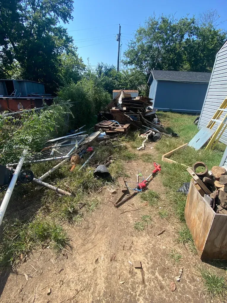 A cluttered outdoor area with scrap metal, wooden debris, a red trimmer, and a blue shed under a clear blue sky.