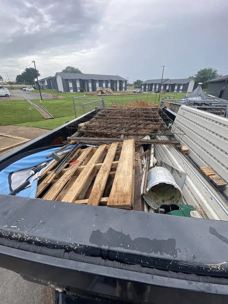 A truck bed filled with wooden pallets, scrap metal, a bucket, and debris, with apartment buildings in the background.