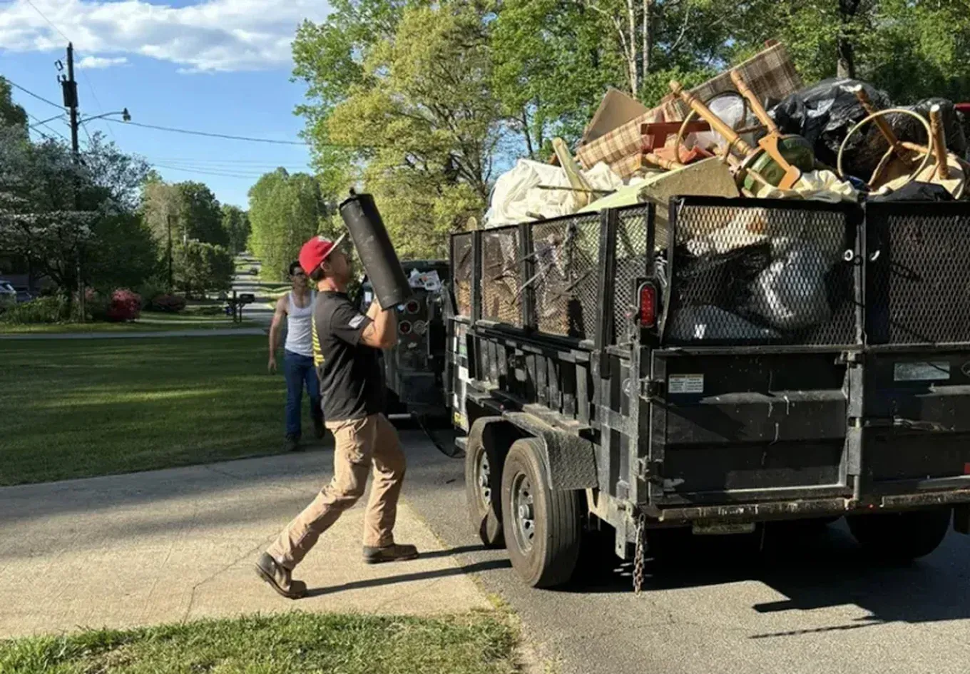 A person in a red cap and work clothes loads a dark cylindrical item into a metal trailer filled with various junk.
