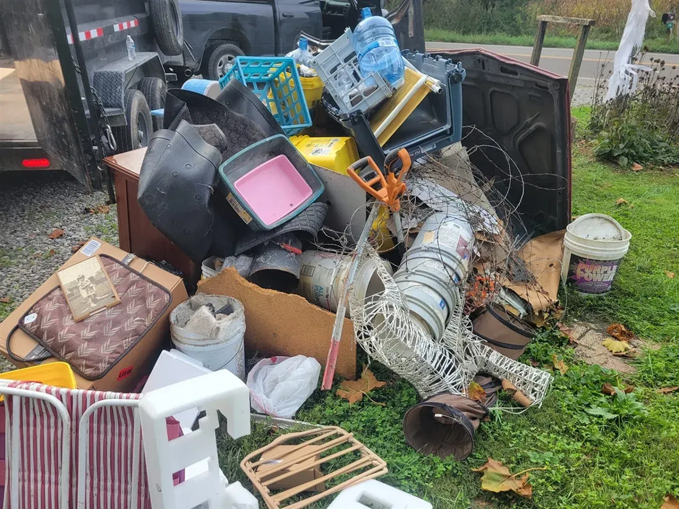 A pile of household junk and debris sits on a grassy patch next to a black trailer.