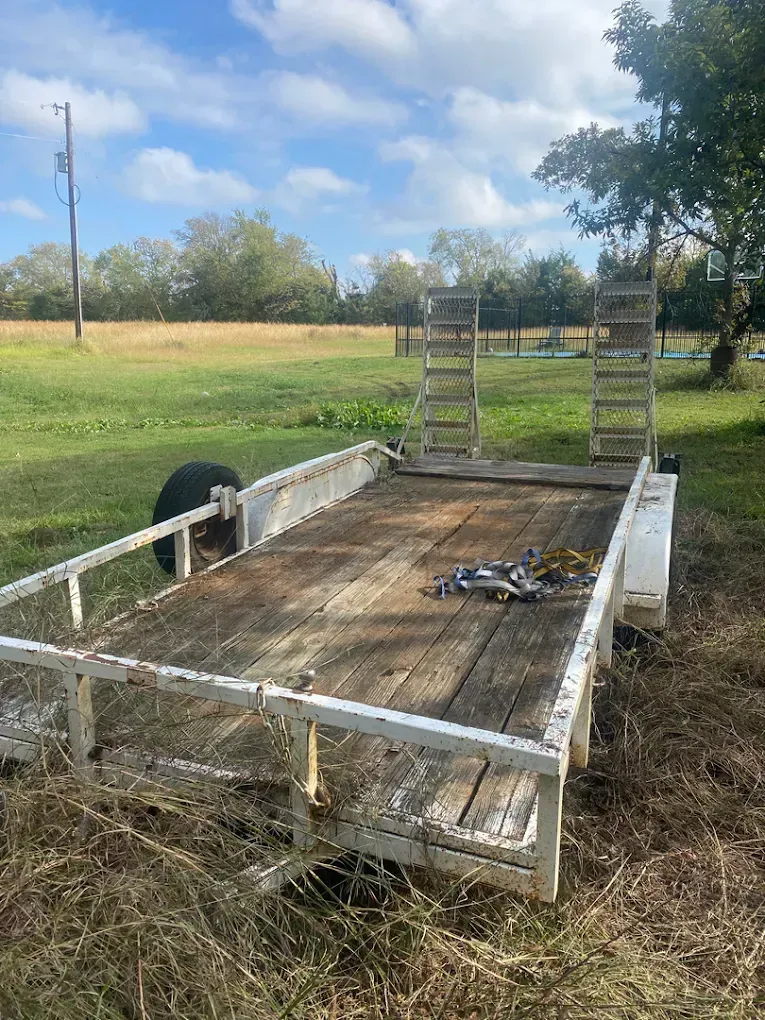 A white utility trailer with a wooden floor and two upright rear loading ramps, parked in a grassy field under a sunny sky.