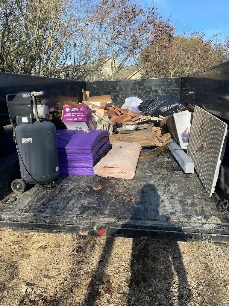 A metal dump trailer filled with miscellaneous debris, including an air compressor, purple foam mats, and scrap wood.