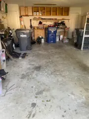 A cluttered garage featuring storage cabinets, shelves, two large trash bins, and miscellaneous items on a concrete floor.
