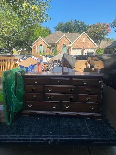 A dark wood dresser with multiple drawers sits in the bed of a truck, surrounded by boxes, parked in front of a house.