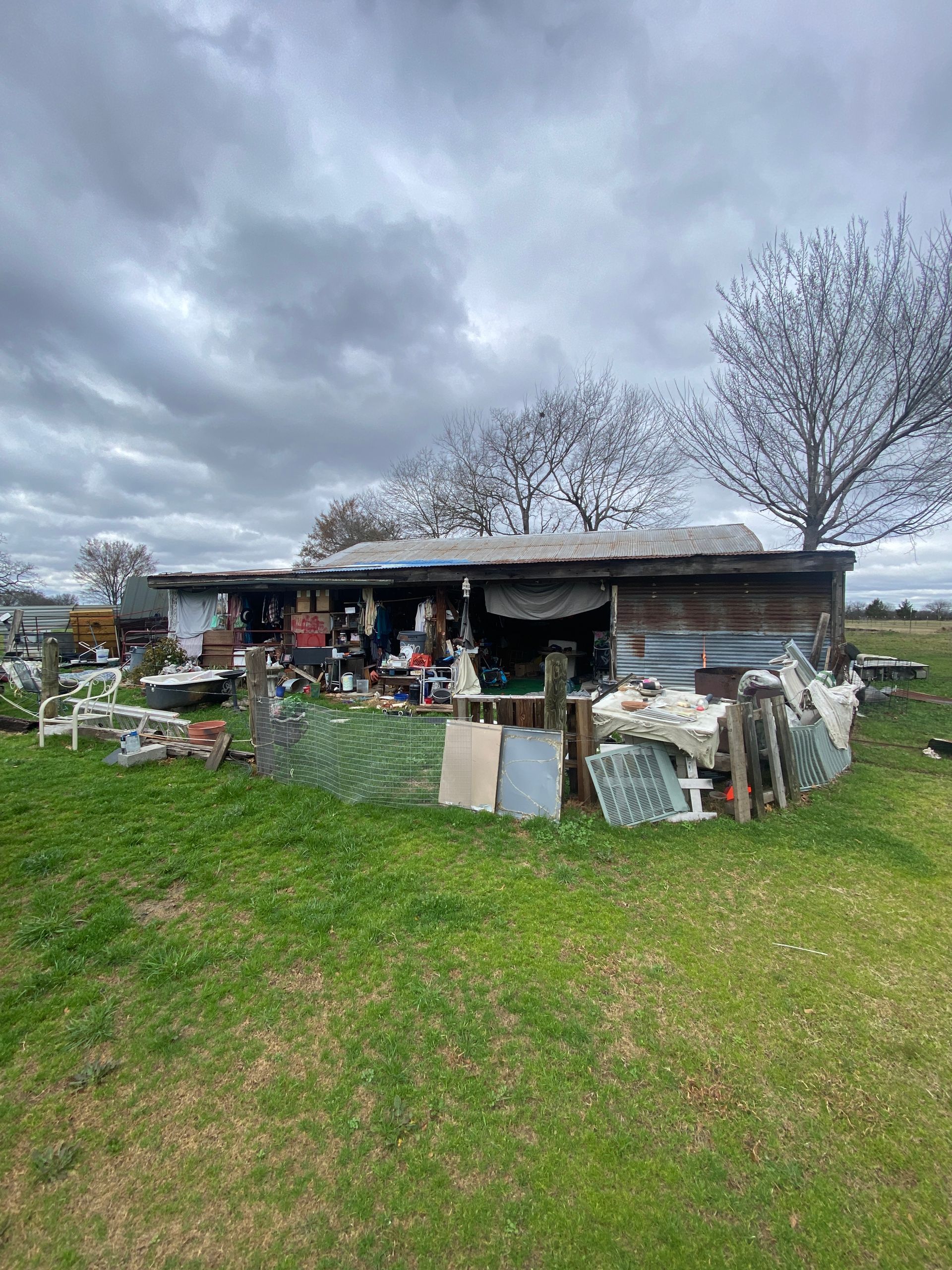 A dilapidated wooden shed in a grassy field, cluttered with various discarded items and makeshift debris.