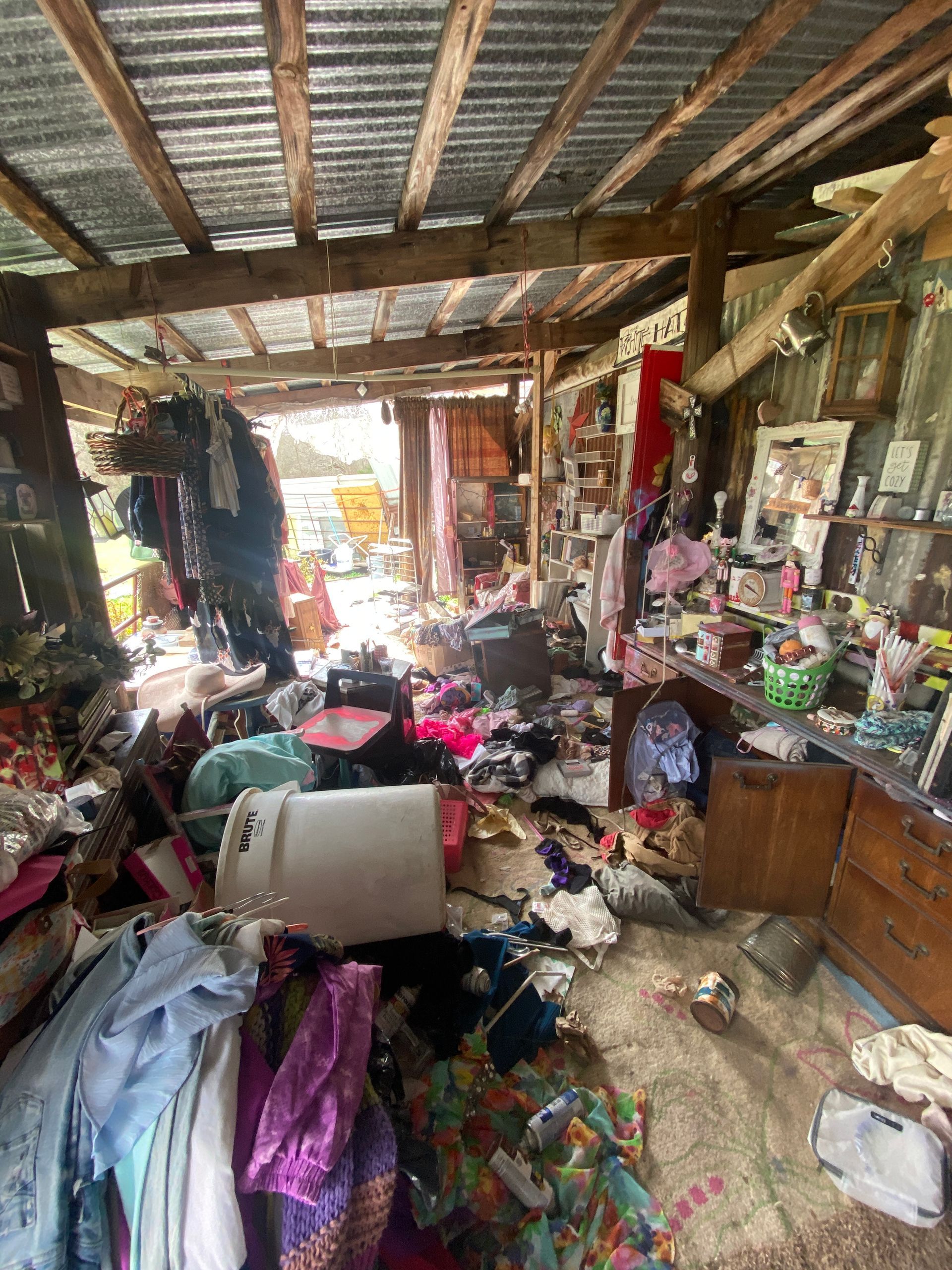 A cluttered, open-walled structure with a corrugated metal roof, filled with scattered clothing, containers, and items.