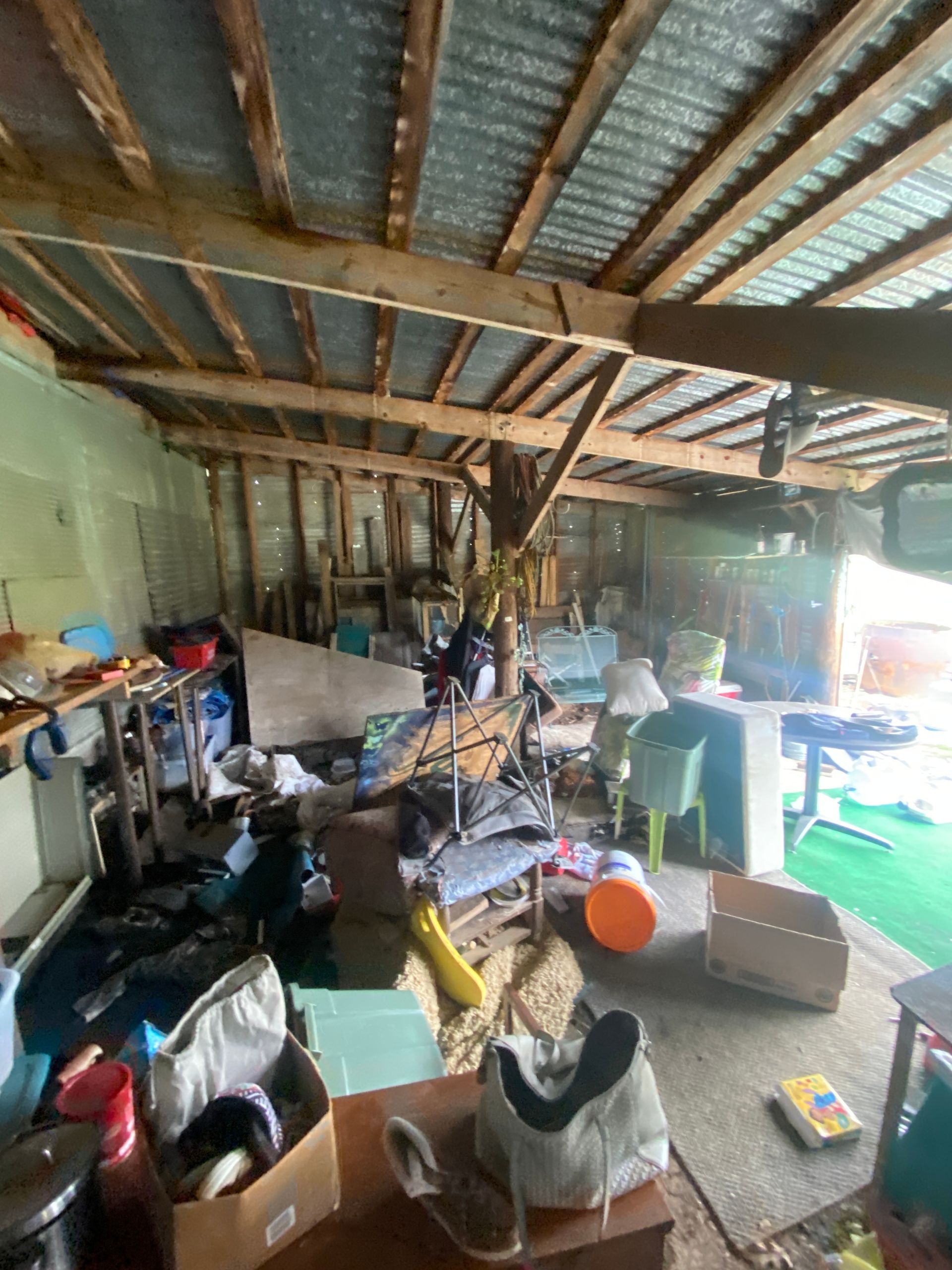A cluttered, open-walled workshop or shed with exposed wooden rafters, corrugated metal roofing, and miscellaneous items.