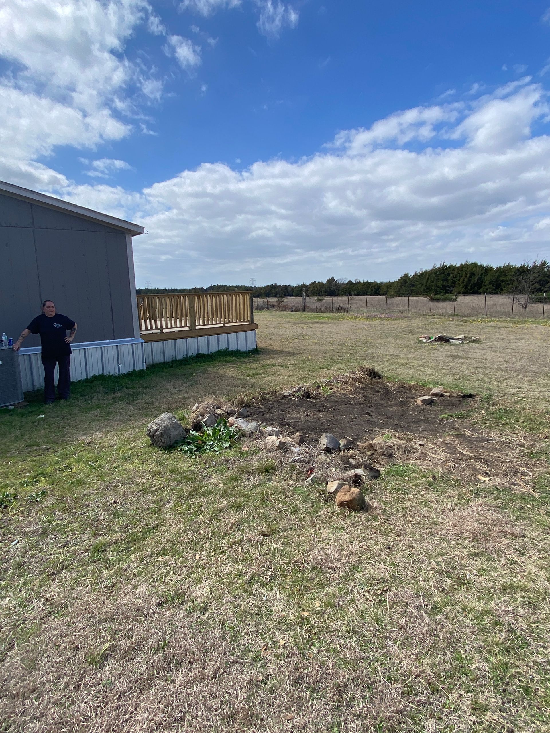 A person stands by the side of a grey house with a wooden deck frame, next to a small, stone-bordered garden patch.