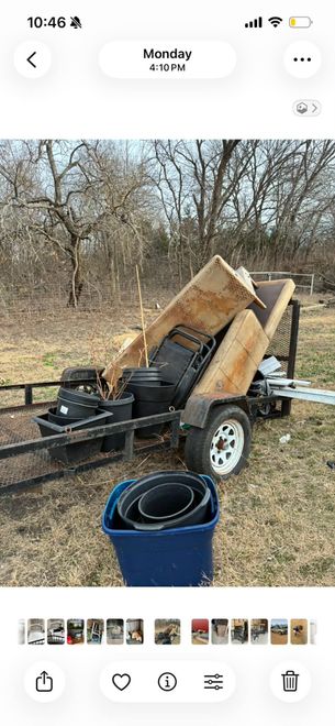 A utility trailer loaded with plywood, black plastic containers, and a blue bin, parked in a wooded area.