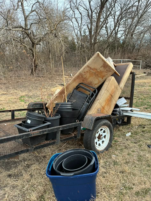 A trailer loaded with old furniture and plastic plant pots sits on dry grass outdoors, with a blue bin in the foreground.