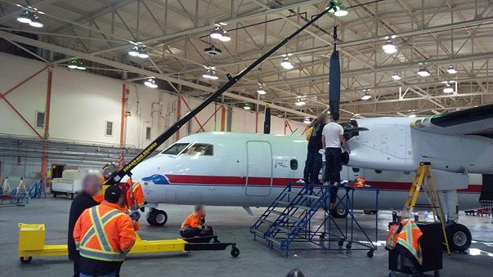 Airplane in a hangar with workers in orange safety gear performing maintenance under the wing.