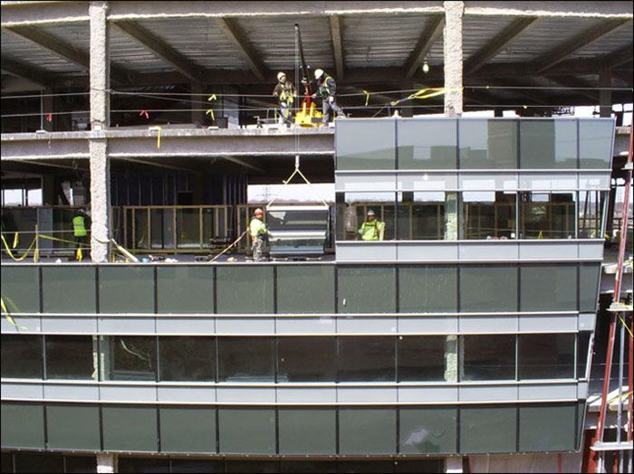 Construction workers on a building facade with dark glass panels under a steel roof.
