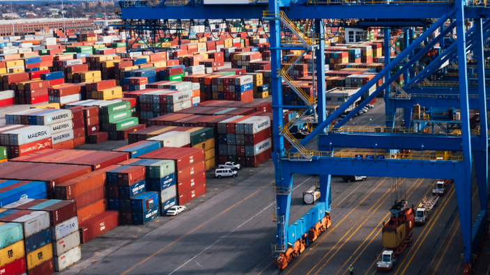 Shipping containers stacked at a busy port beside blue cranes and cargo trucks