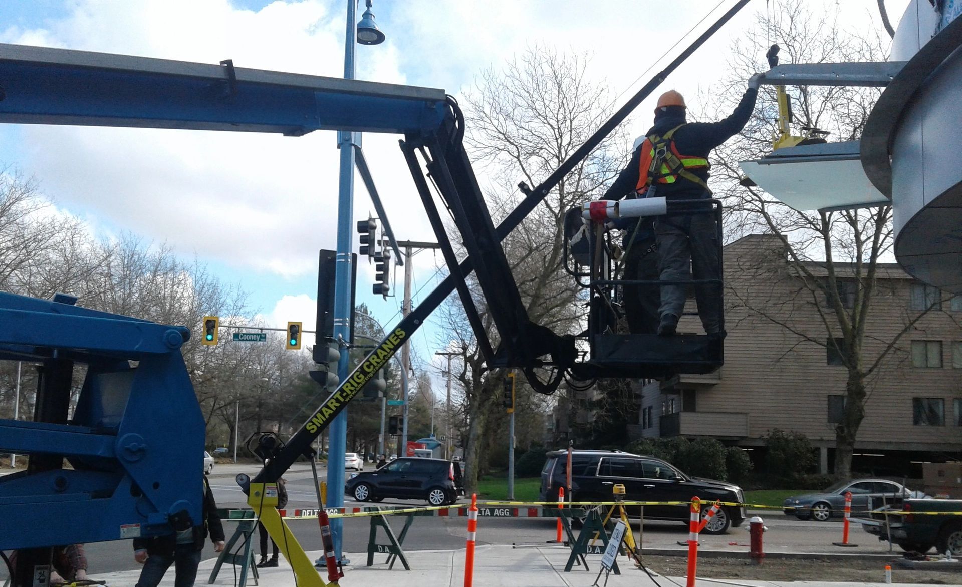Workers in orange vests on blue lifts trimming tree branches by a street