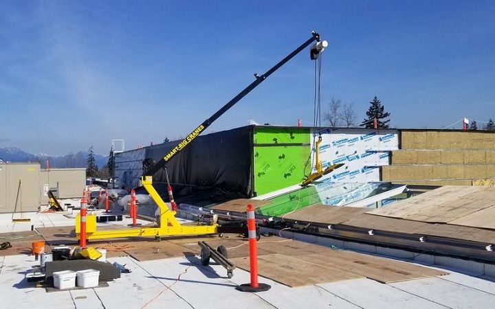 Construction crane lifting materials on a flat rooftop, with orange cones and wrapped panels nearby.