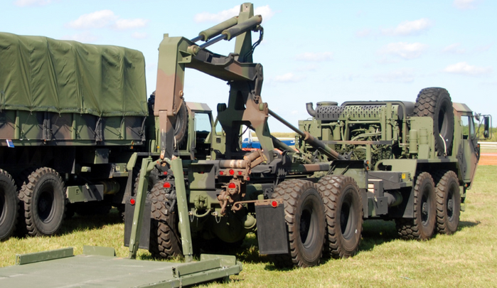 Military trucks with a crane trailer parked on grass under a blue sky