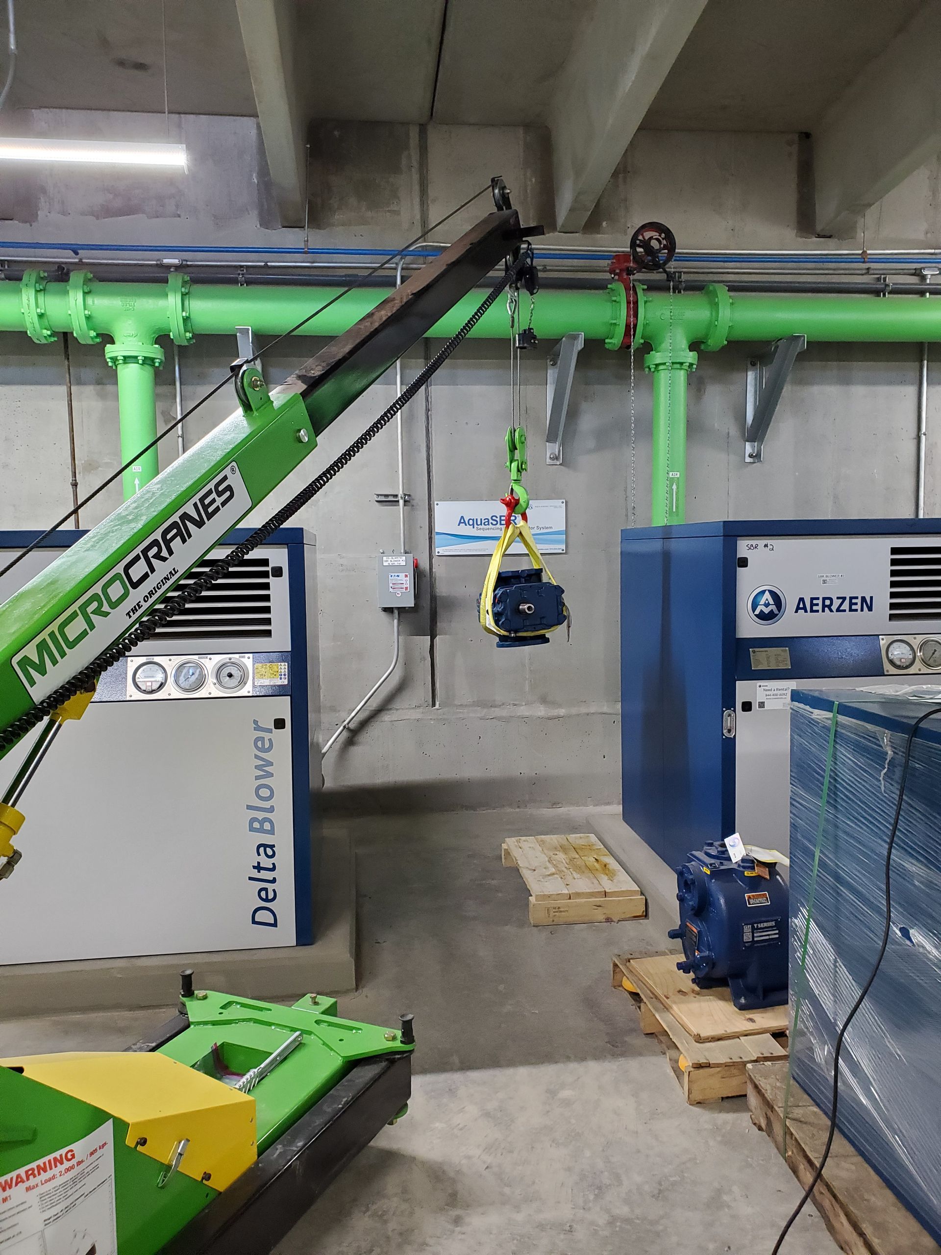 Green industrial machinery and pipes in a factory room with a blue machine and conveyor arms.
