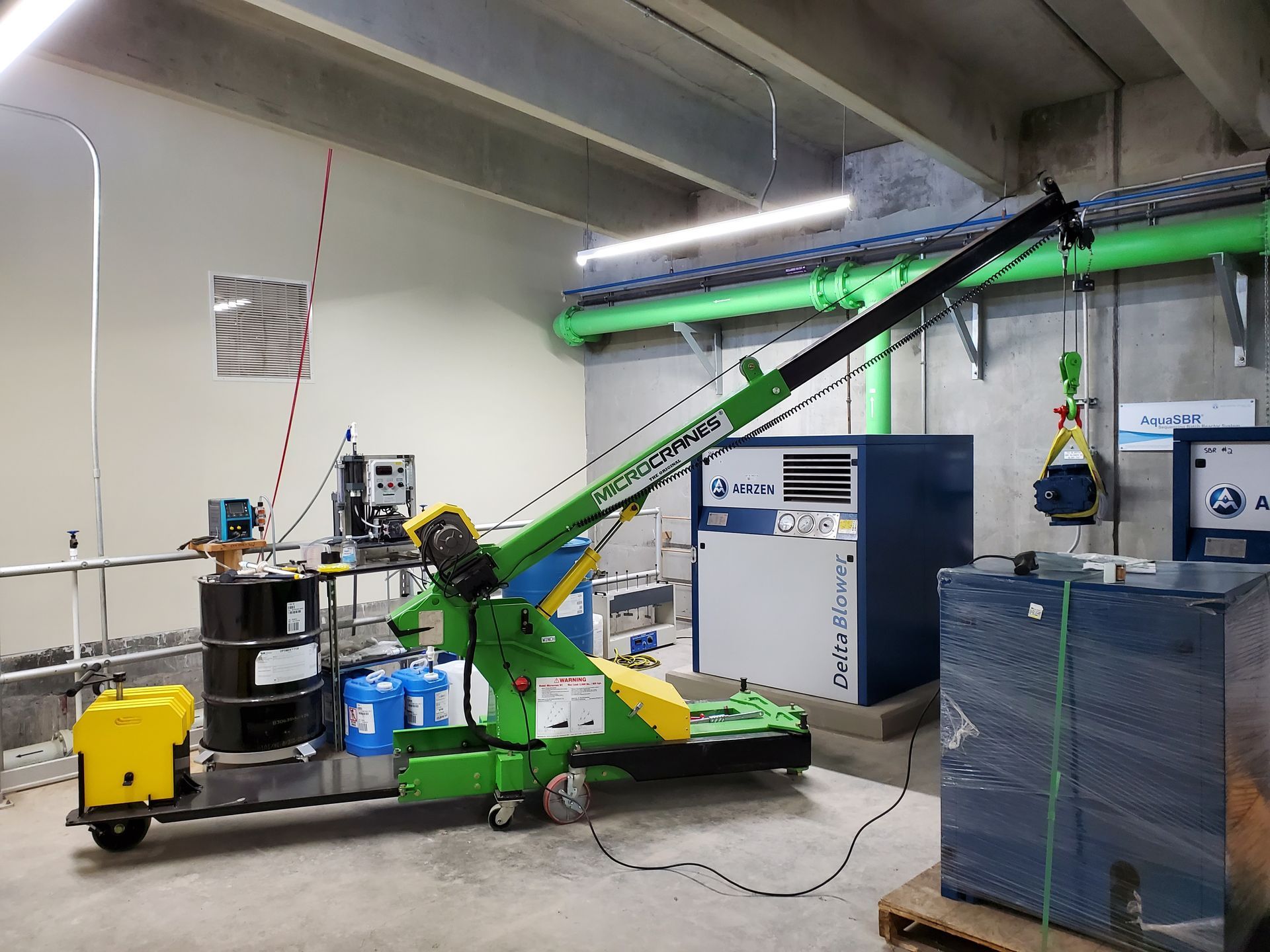 Green industrial machine with long conveyor arm in a warehouse, beside blue equipment and a yellow bin