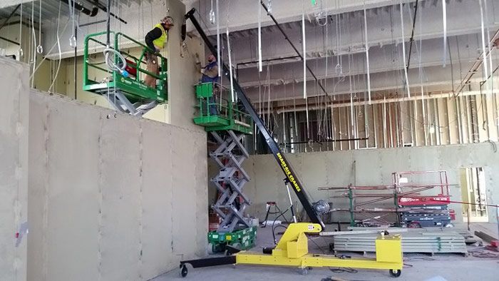 Construction site with a yellow scissor lift raising workers near a wall under a ceiling of hanging wires and pipes