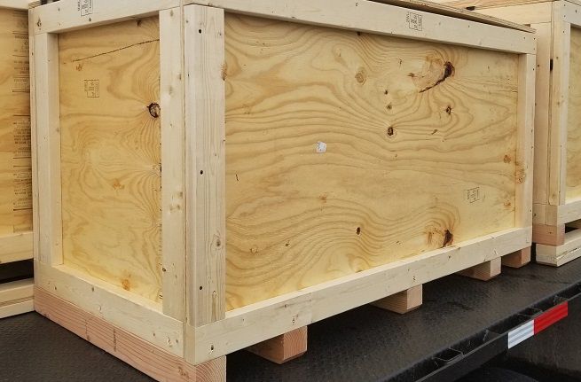 Wooden shipping crate secured on a truck bed, with pale plywood panels and corner framing.