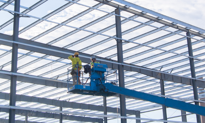 Two workers in safety gear on a blue boom lift assembling a steel-framed roof structure