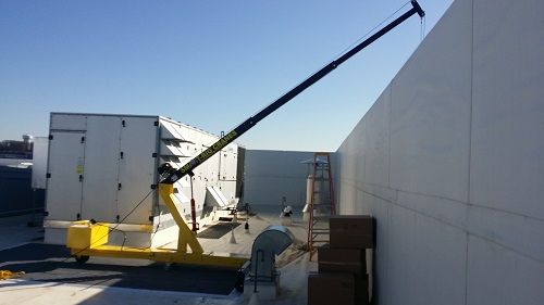 Rooftop crane arm angled over a white wall, with a worker near equipment in a bright industrial area