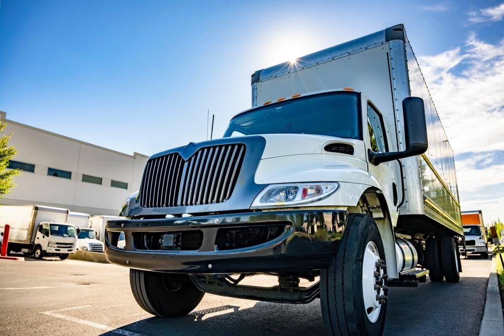 White and black box truck in a parking lot, sunny day, other trucks and a building in the background.