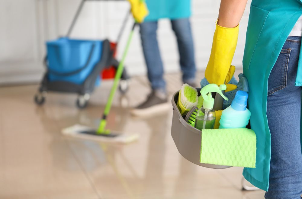 A Woman is Holding a Bucket of Cleaning Supplies in a Room — Complete Clean Home Cleaning in Woonona, NSW