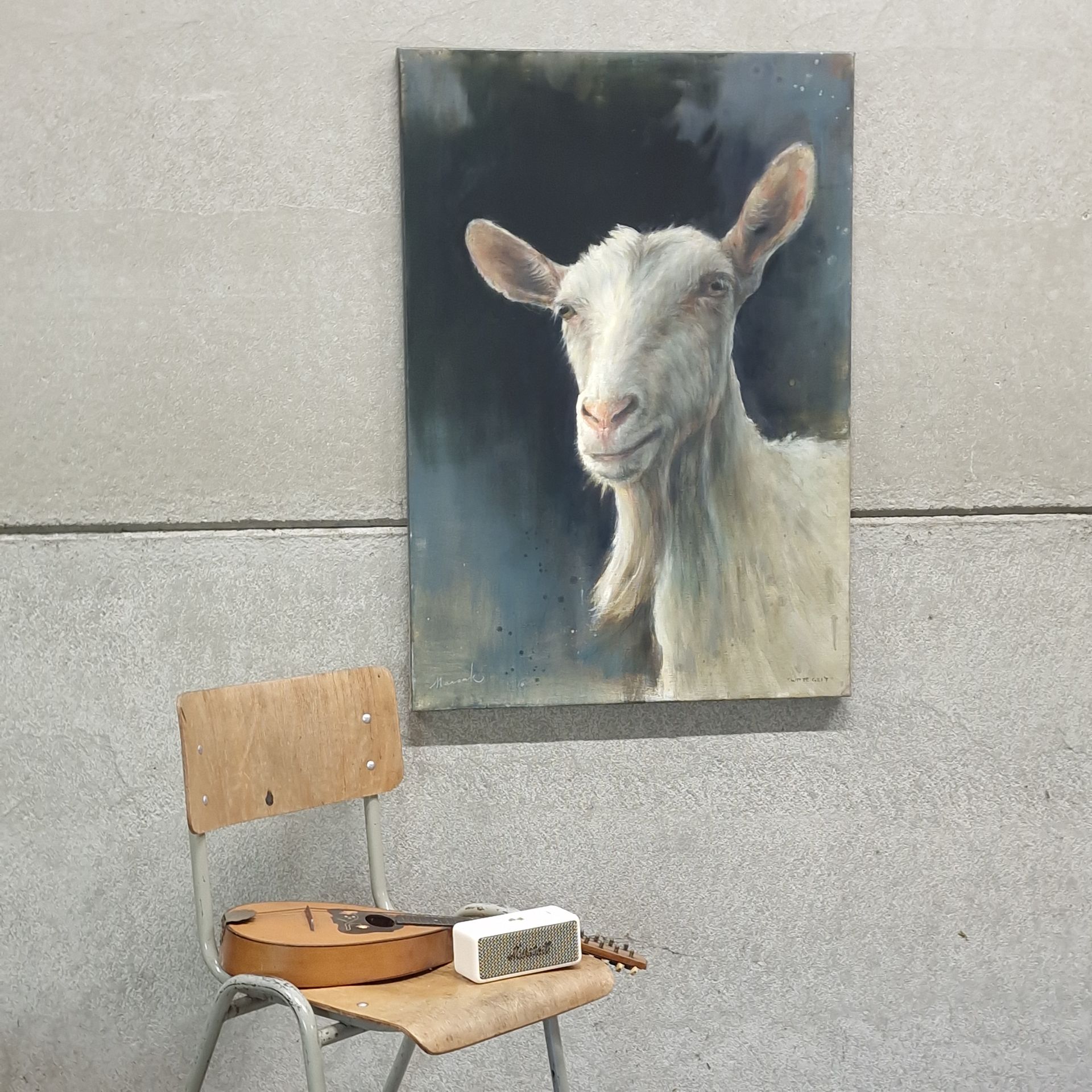 Schilderij van een geitenportret op een grijze muur boven een houten stoel met een snaarinstrument en een klein doosje.