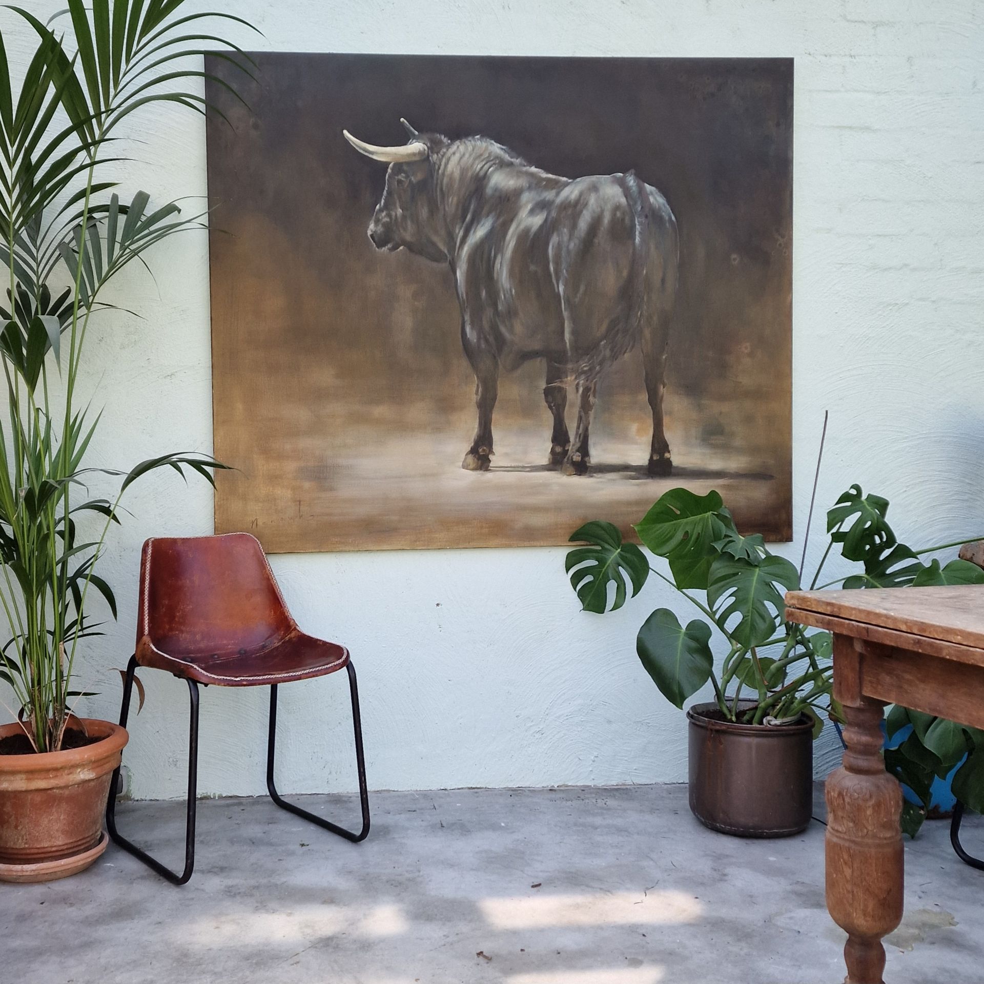 Een terras met een schilderij van een stier aan de muur, een rode stoel, potplanten en een houten tafel.